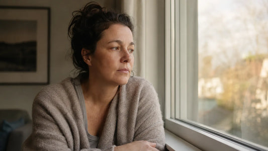 A woman in her mid-40s sitting by a window in the early morning, looking out quietly.