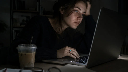 A woman working at a laptop at night, one hand wearing a turquoise ring, face lit by the screen.