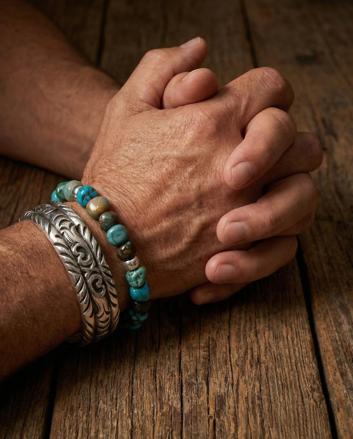 Clasped hands resting on a wooden table wearing a grounding Tibetan turquoise bead bracelet and an oxidized silver cuff.
