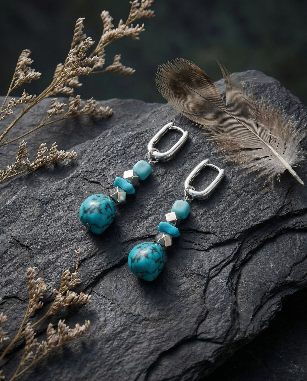 Silver Tibetan turquoise drop earrings elegantly displayed on dark rugged slate stone alongside a natural feather.