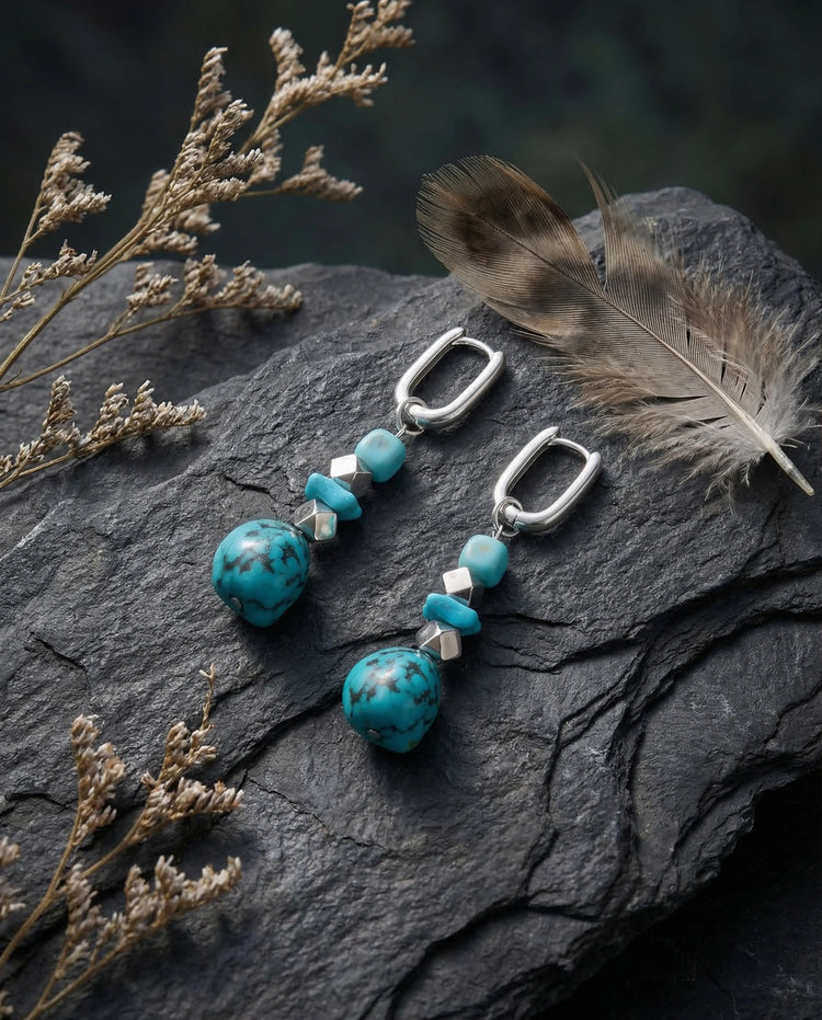 Silver Tibetan turquoise drop earrings elegantly displayed on dark rugged slate stone alongside a natural feather.