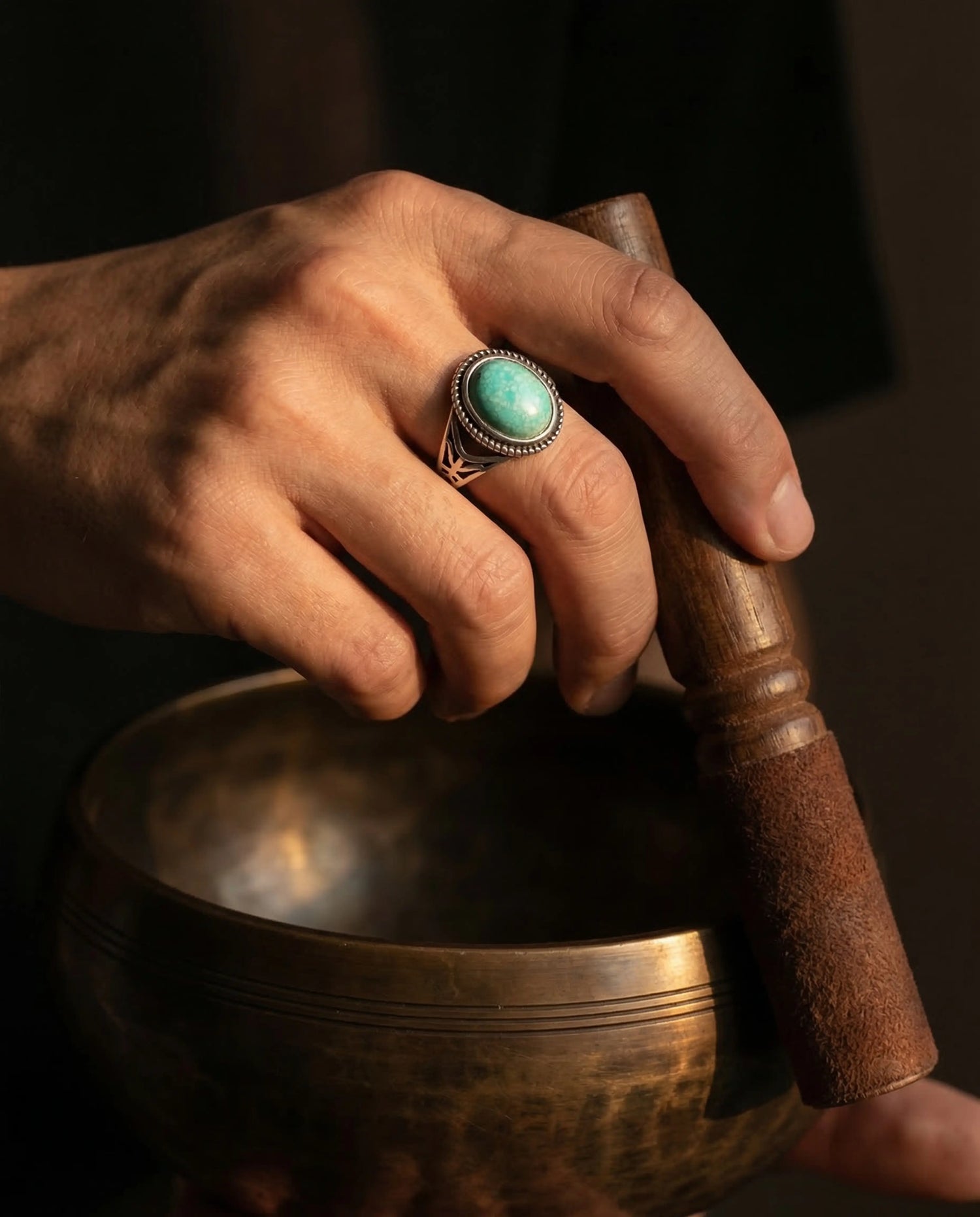 A hand wearing an oxidized silver Tibetan turquoise signet ring holding a mallet over an antique singing bowl during meditation.