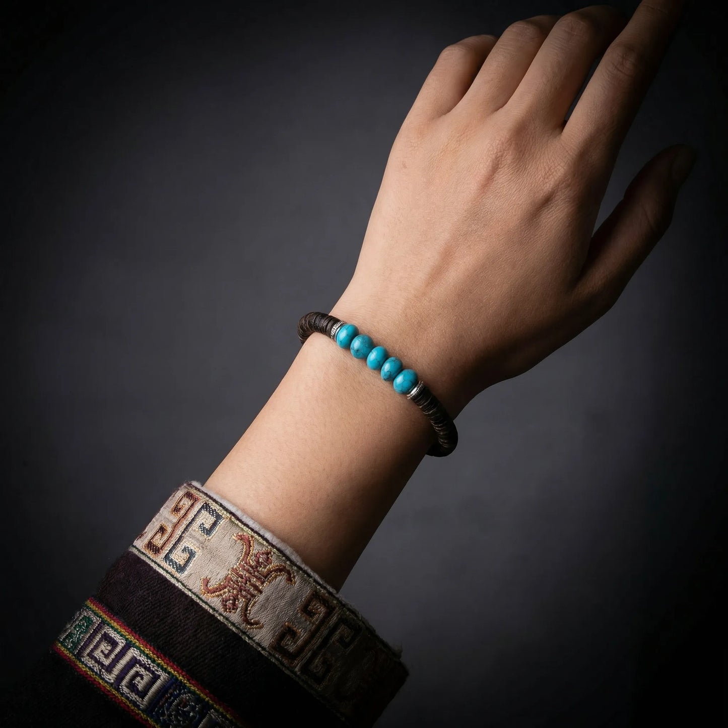 A hand reaching upward wearing the grounding Tibetan turquoise bracelet against a dark, moody background.