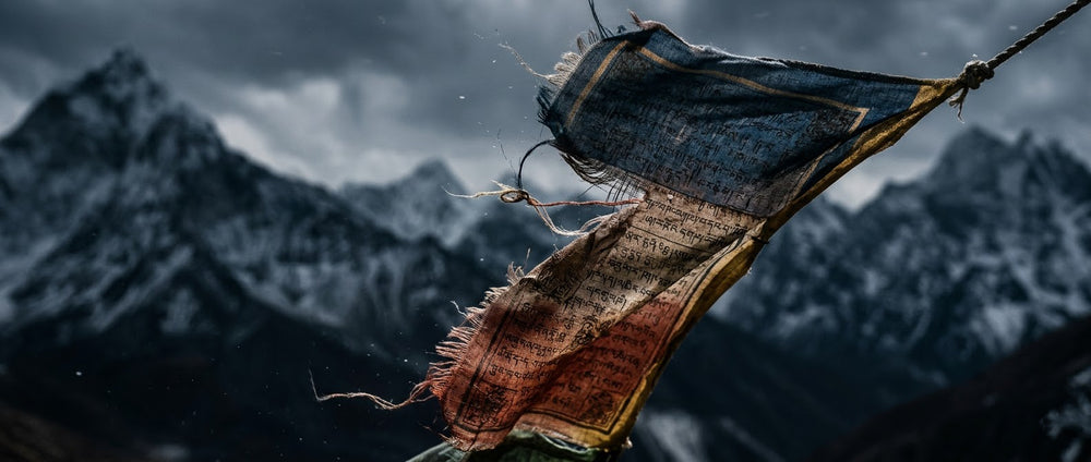 A dramatic twilight view of a sacred Himalayan peak, with blurred Tibetan prayer flags fluttering in the foreground. This majestic landscape represents the spiritual origins and the untamed, raw energy that inspires every handcrafted piece of Tashi Turquoise jewelry.