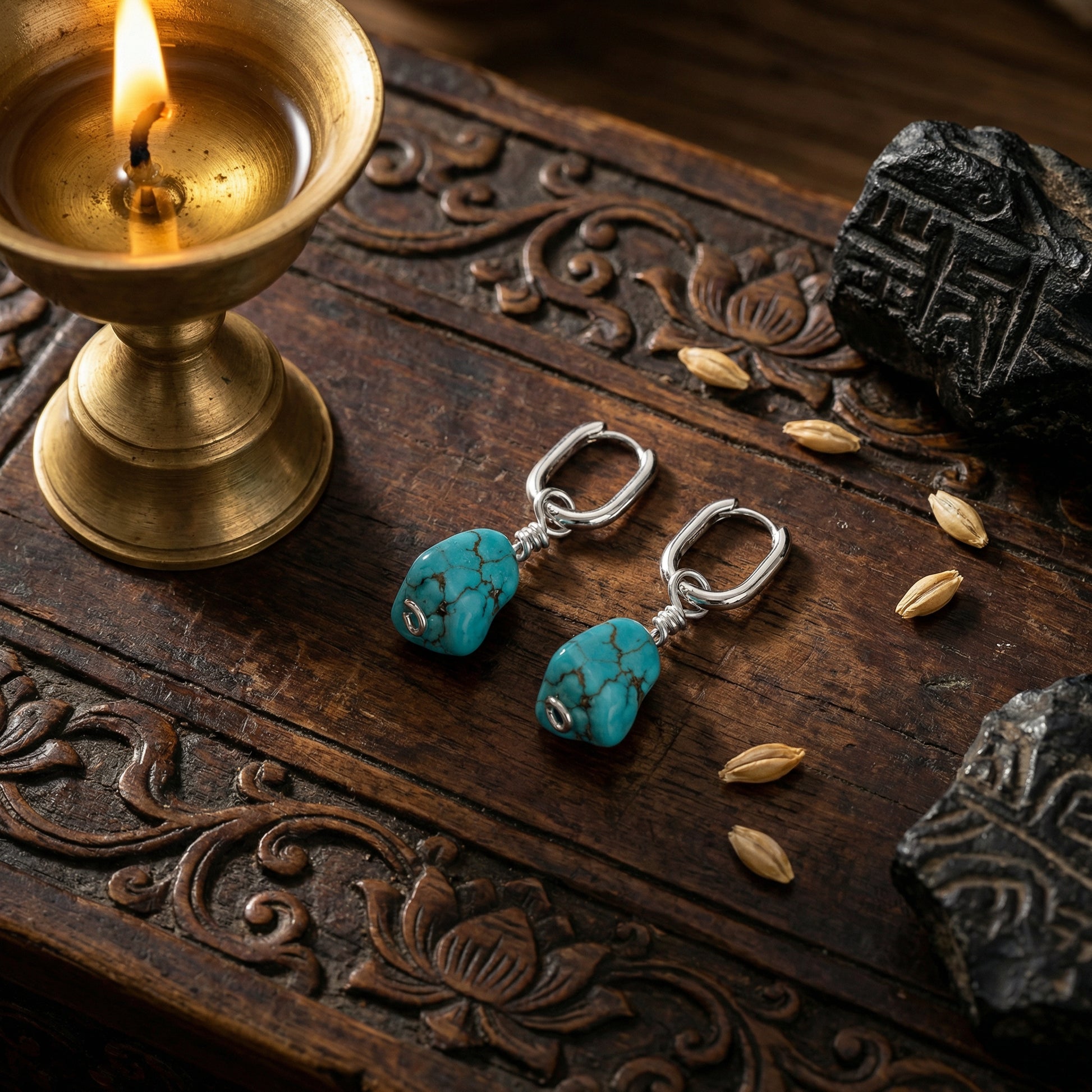 The Raw Drop earrings displayed on a carved wooden box with a lit brass candle, reflecting Himalayan heritage.