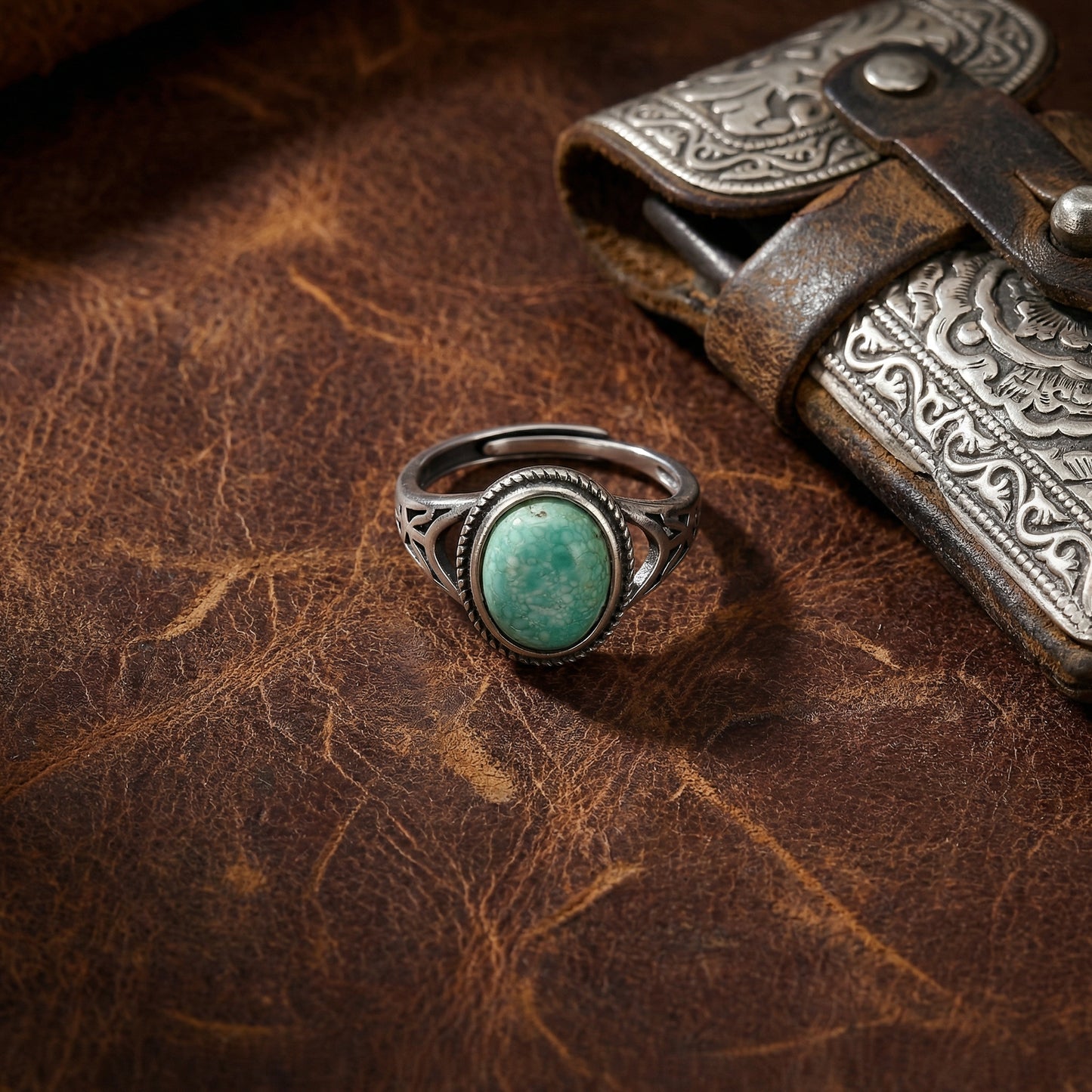 The Nomad's Shield Ring displayed on a rustic leather surface near a vintage silver piece.